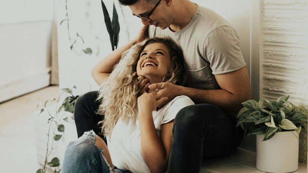 A happy couple sitting indoors, smiling affectionately while leaning against each other.