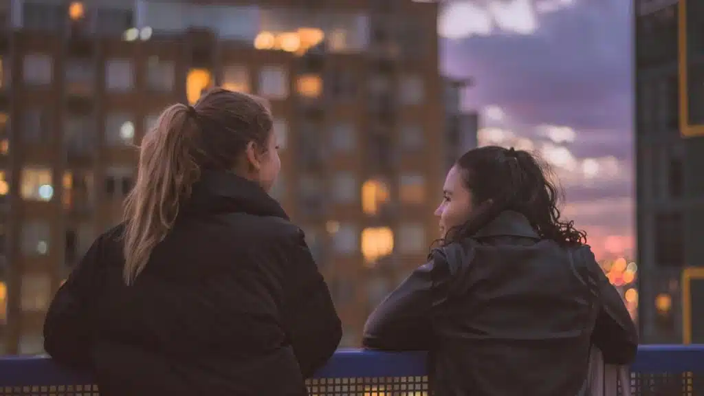 A pair of women leaning on a railing and talking as city lights glow at dusk.