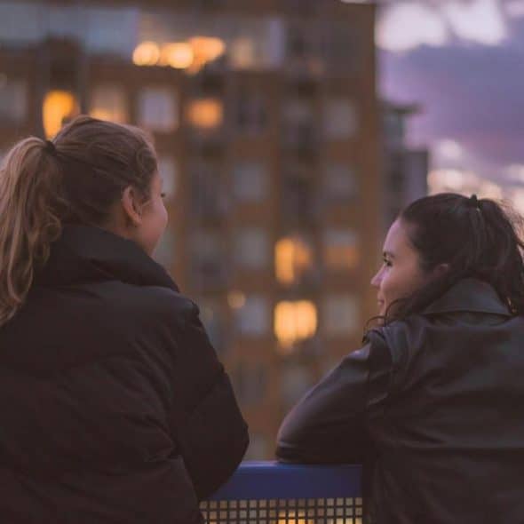 A pair of women leaning on a railing and talking as city lights glow at dusk.