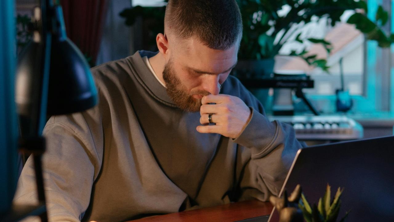 A bearded man concentrating at a desk while working on a laptop indoors.