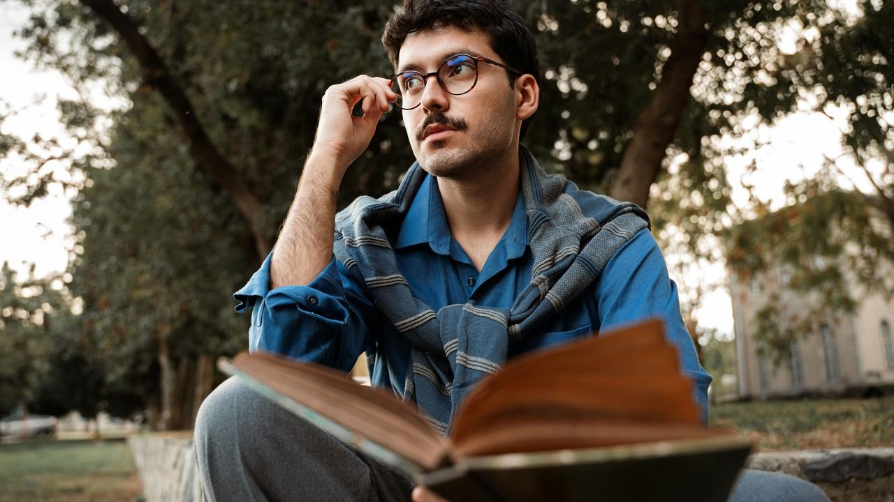 A man adjusting his glasses while reading a book outdoors.
