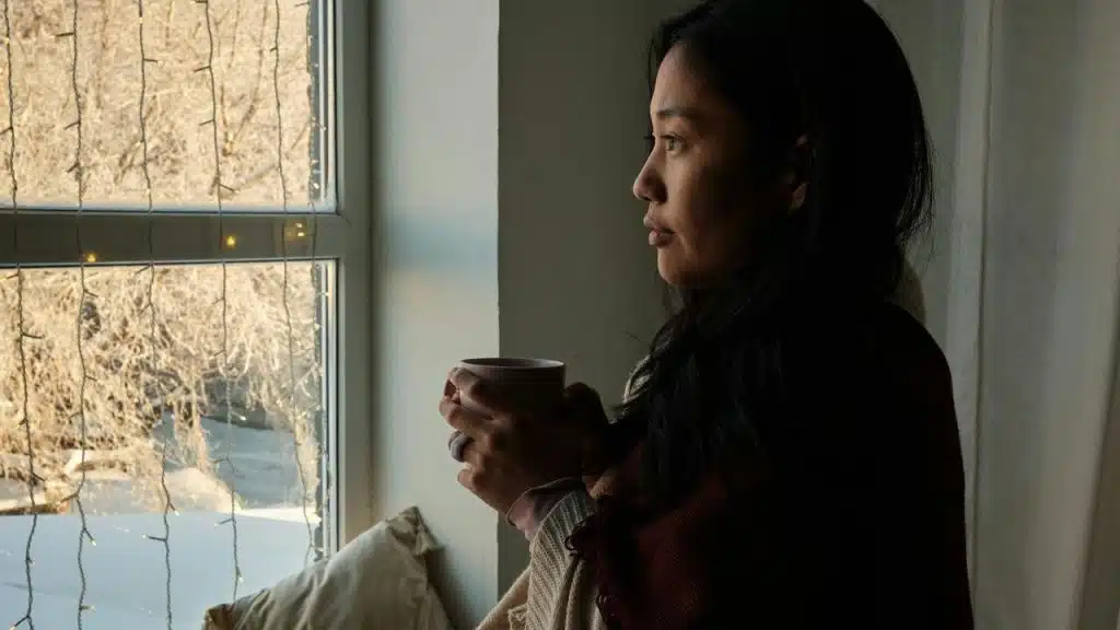 A woman holding a mug while looking out a window with winter scenery and string lights.