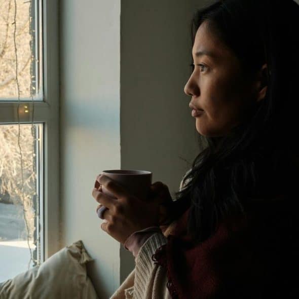 A woman holding a mug while looking out a window with winter scenery and string lights.