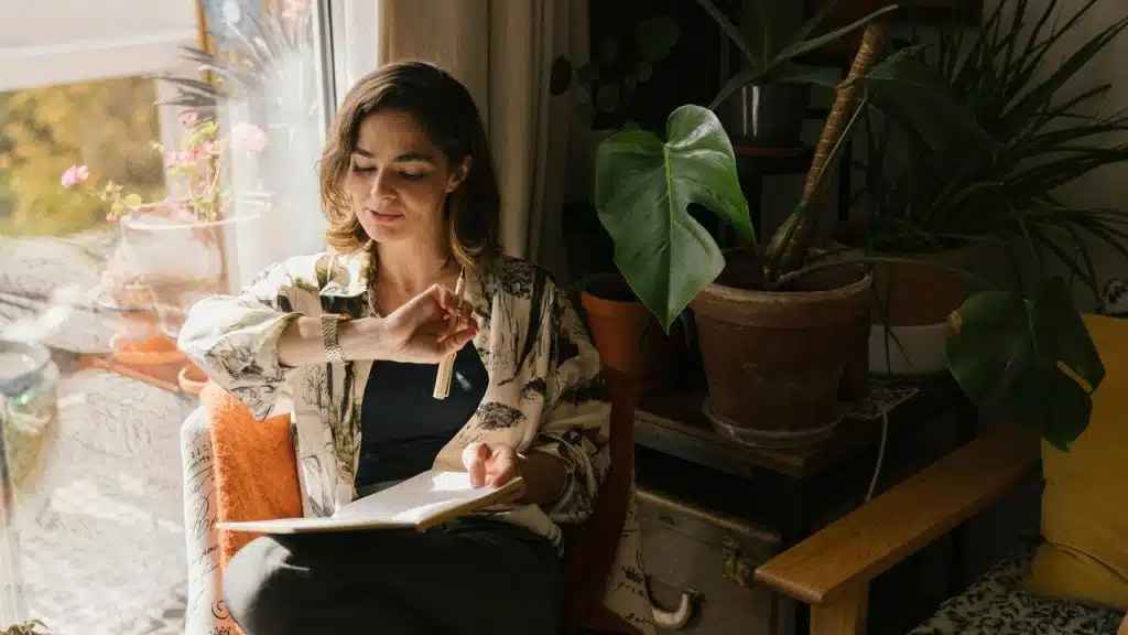 A woman sitting by a window, writing in a notebook surrounded by houseplants.
