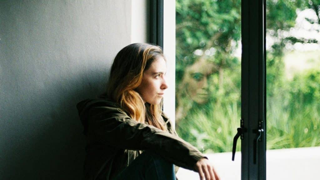 A woman sitting by a window, looking outside with her reflection visible in the glass.