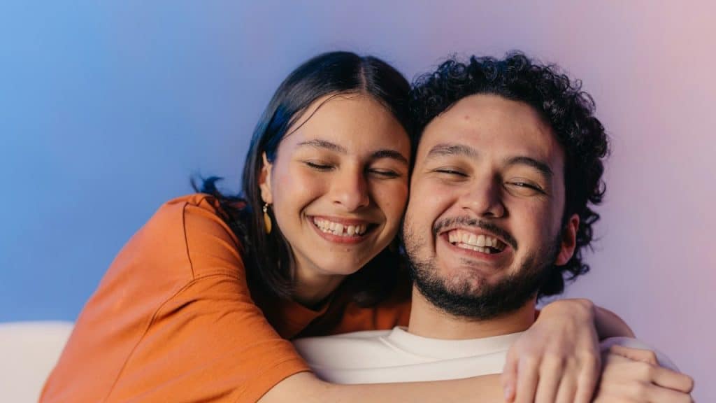 A woman holding a bearded man close as they smile together while sitting on a couch.