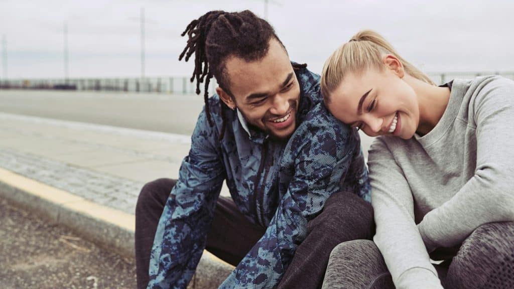 A smiling woman leaning on a smiling man’s shoulder as they sit outside on a pavement.