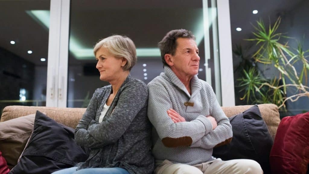An older couple sits back-to-back on a couch with their arms crossed, looking away.