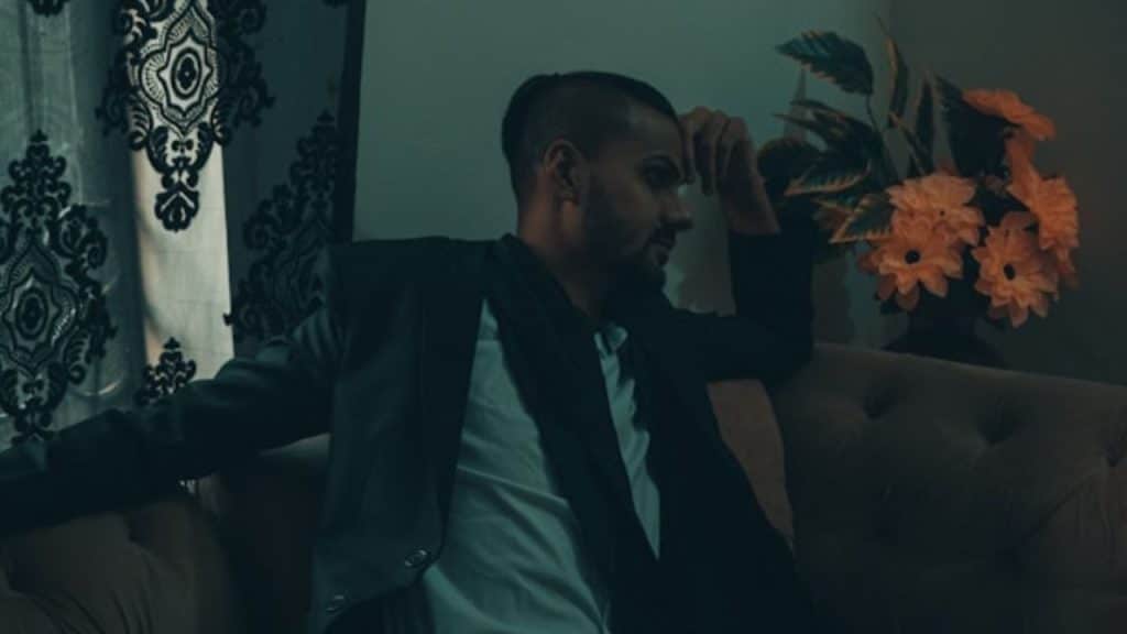 A man in a dark blazer sits on a sofa next to orange flowers and patterned curtains.