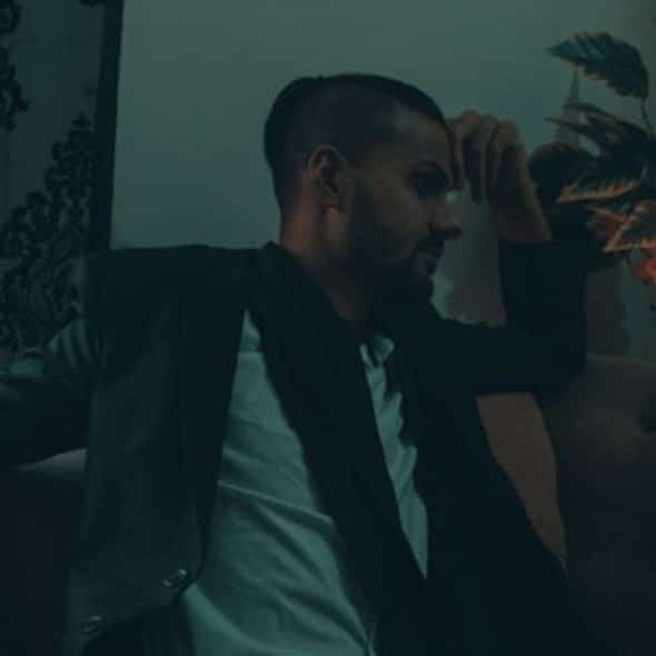 A man in a dark blazer sits on a sofa next to orange flowers and patterned curtains.