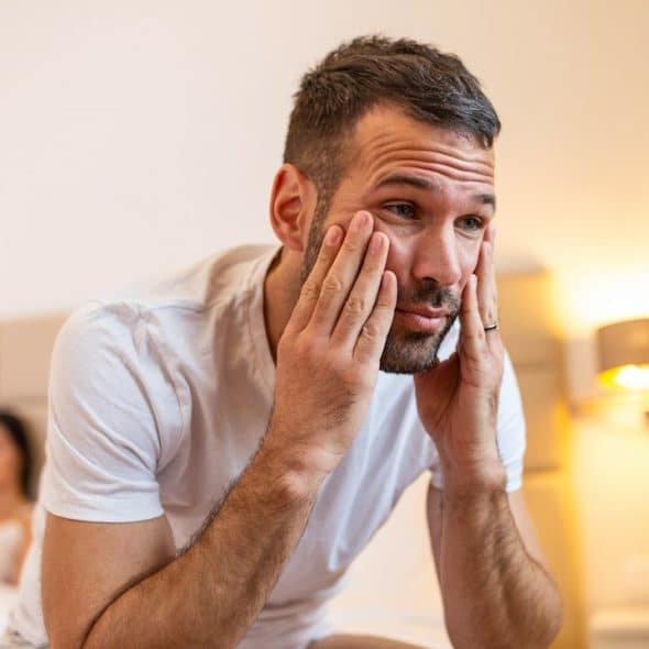 An upset, lightly bearded man holding his face in his hands while his wife lies in a bed behind him.
