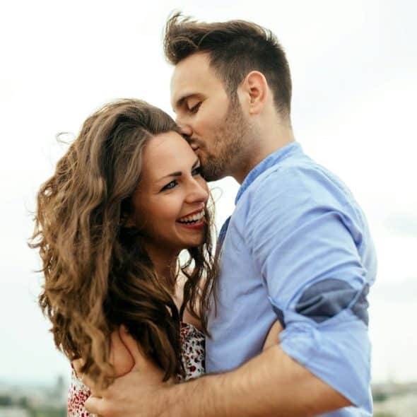 A lightly bearded man kissing a smiling woman on her temple.