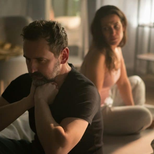 A bearded man holding his chin with his hands, sitting with his back to an upset woman, looking at him.