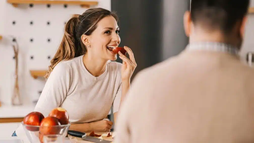 A woman smiling while eating an apple in a kitchen, with another person nearby.
