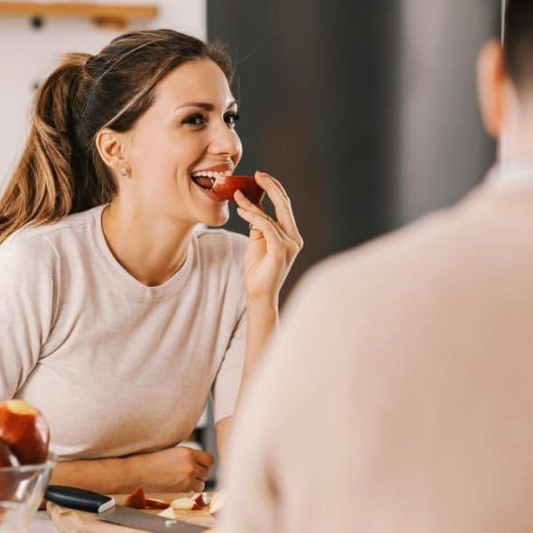 A woman smiling while eating an apple in a kitchen, with another person nearby.