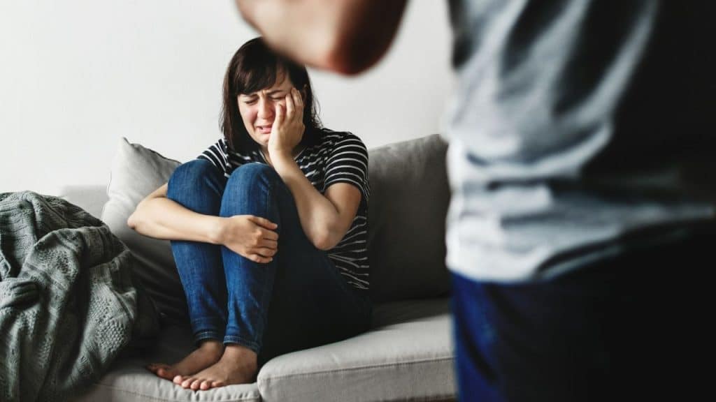 A woman sits crying on a couch while a blurred figure stands in the foreground.