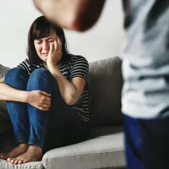 A woman sits crying on a couch while a blurred figure stands in the foreground.