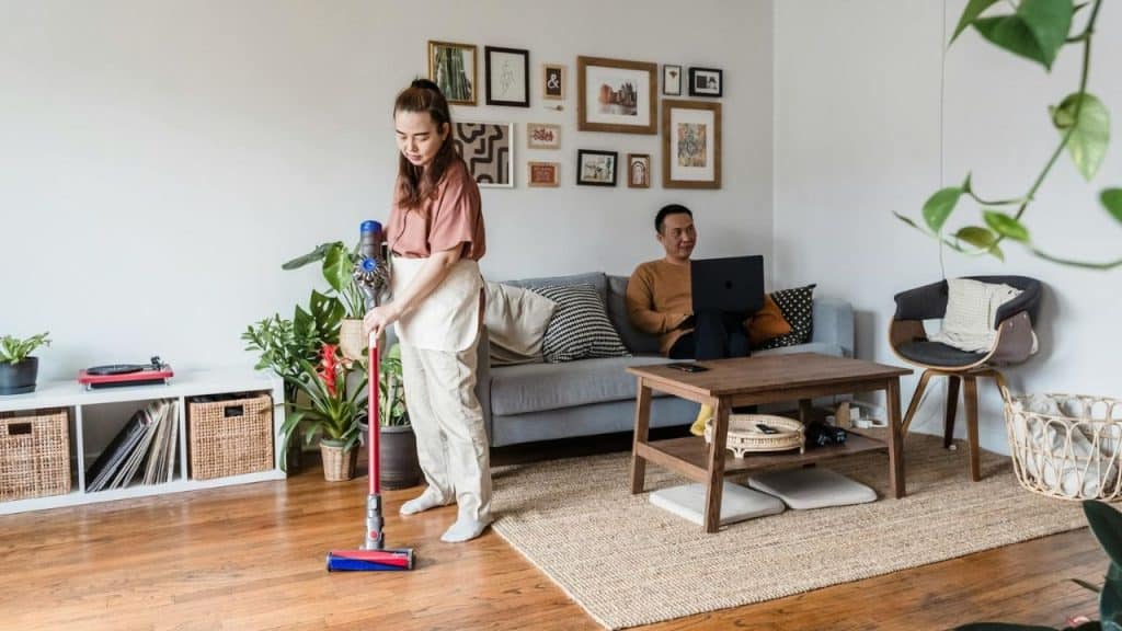 A woman vacuums a wooden floor while a man sits on a sofa using a laptop.