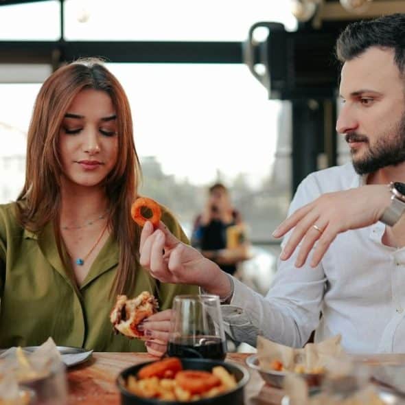 A man offers a fried onion ring to a woman holding a partially eaten burger.