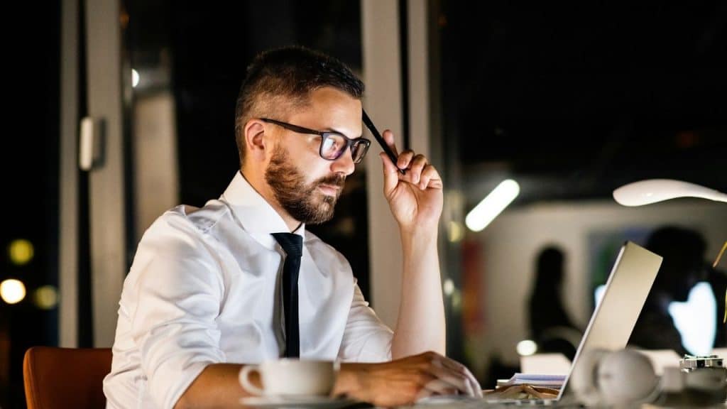 A man in glasses and a tie working late at a laptop in a dark office.