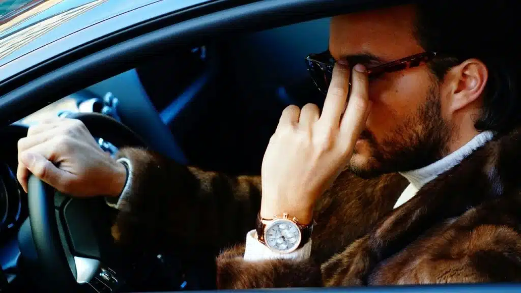 A man wearing a fur coat and watch holds a steering wheel and adjusts sunglasses.