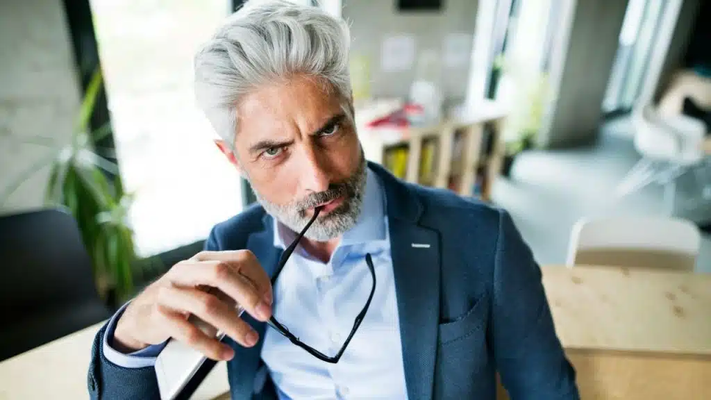 A middle-aged man with gray hair and a beard, wearing a suit, holding black glasses.