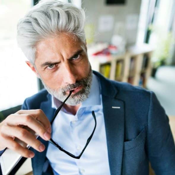 A middle-aged man with gray hair and a beard, wearing a suit, holding black glasses.