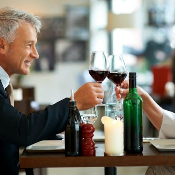 A mature couple making a toast by clinking glasses while sitting at a table at a restaurant.