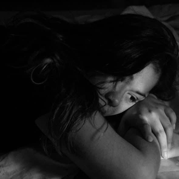 A black and white image of a woman lying on a bed with her head resting on her arms, looking sad or troubled.