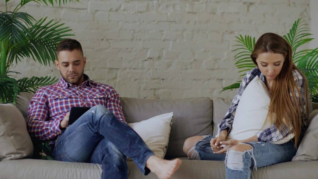 A couple is sitting separated on a sofa, both looking down and engrossed in their own activities