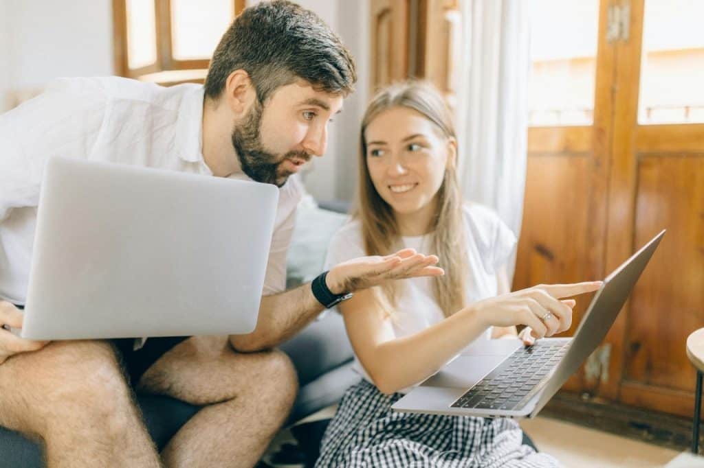 A man holding a laptop on his lap while looking at his wife’s laptop as she shows something on it to him.