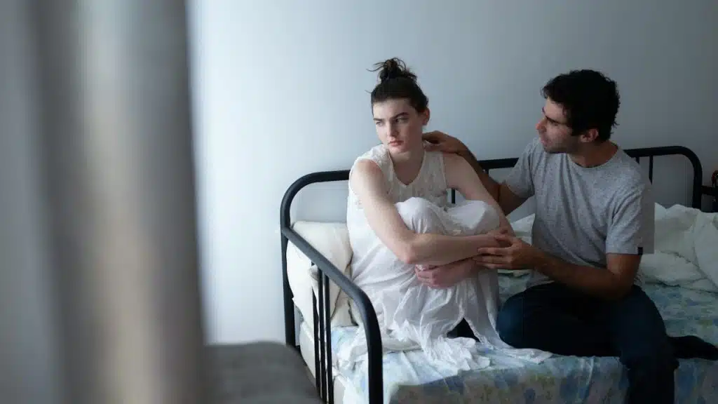 A woman looking to the side as a man asks her about something while they sit on a bed.