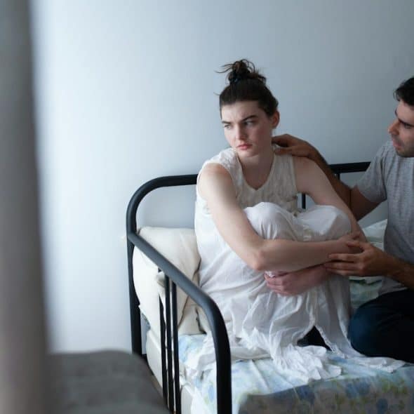 A woman looking to the side as a man asks her about something while they sit on a bed.