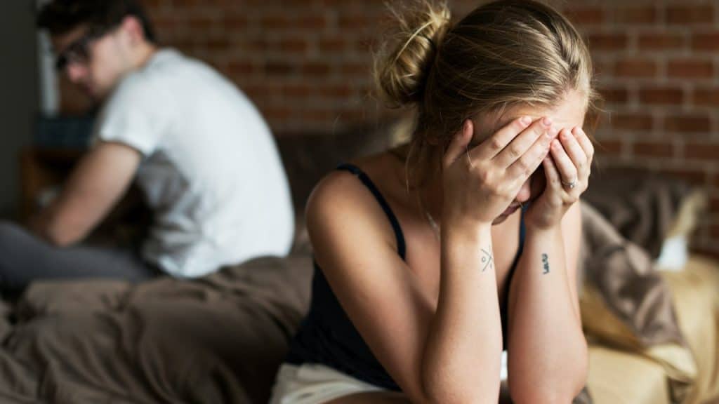 A couple sitting on opposite sides of a bed while a woman cries with her face hidden in her hands and the man looks at her from the side of his vision.