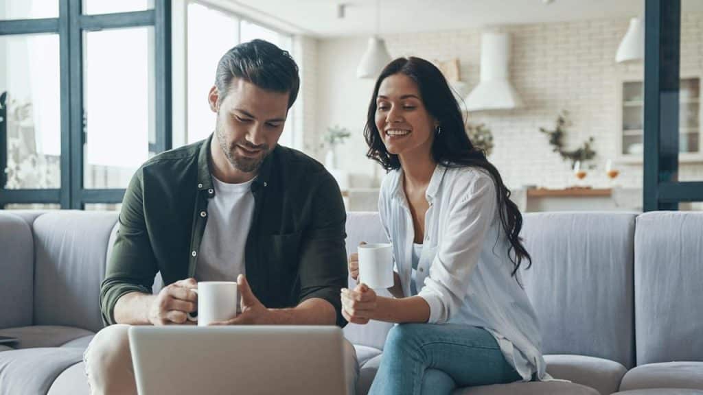 A beautiful young couple drinking coffee and looking at the laptop while sitting on the sofa at home.