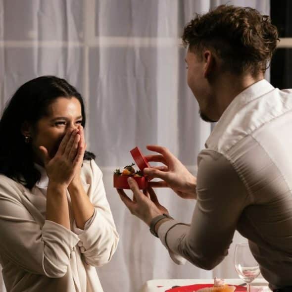 A man opens a small red box for a woman who is covering her mouth.