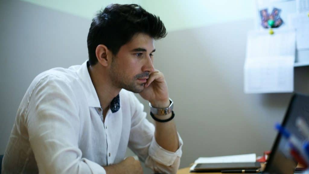 A man with dark hair leans his chin on his hand while looking at a screen.