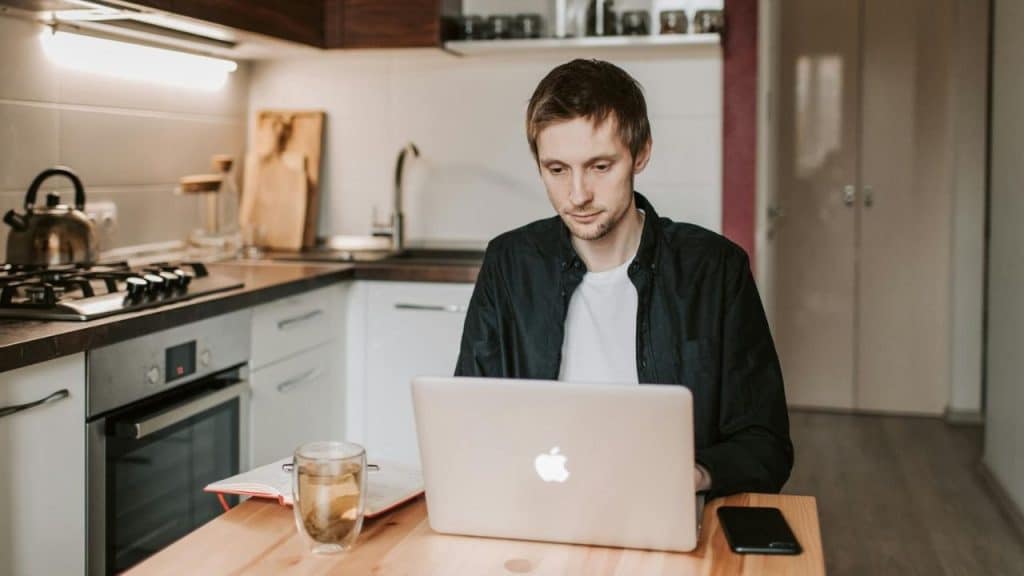 A man sits at a kitchen table using a laptop next to a glass of tea.