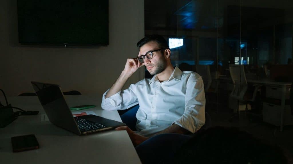 A man wearing glasses sits at a desk in a dark office, looking at a laptop.
