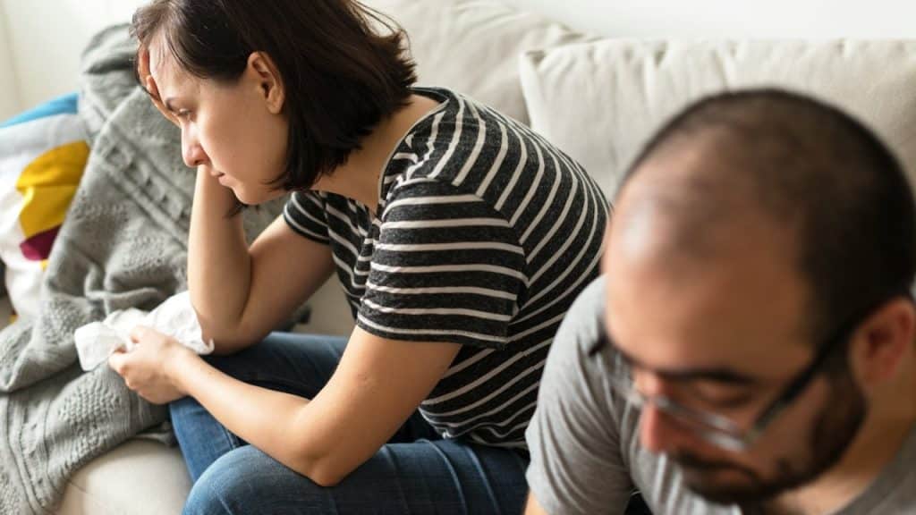 A woman looking distressed while a man looks down as they sit on a couch.
