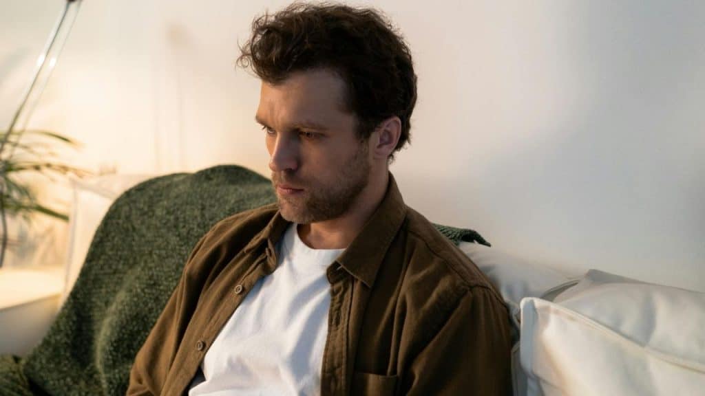 A man with curly hair and a brown shirt sits on a white couch looking down.