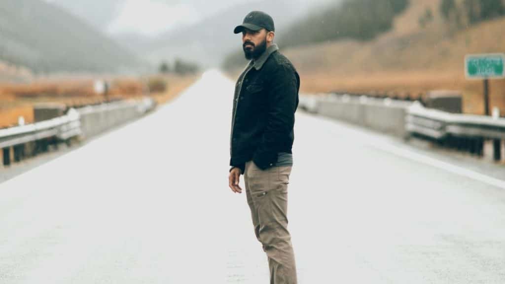 A bearded man wearing a cap and jacket stands on a long, empty, paved road.
