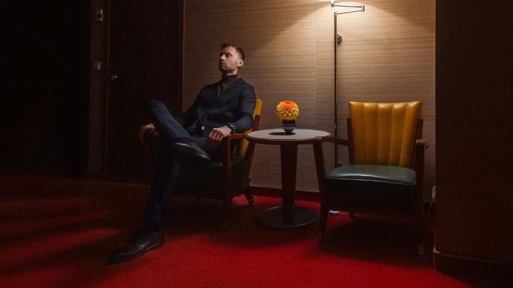 A man sits alone in a chair in a dimly lit room with red carpet.