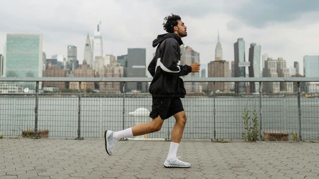 A man in athletic wear runs along a waterfront path with a city skyline behind him.