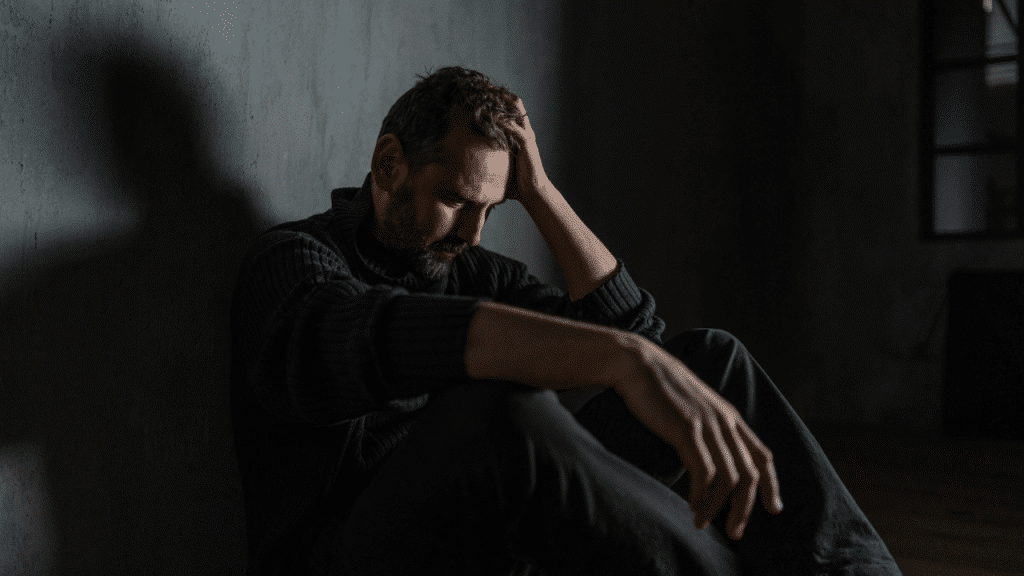A man sits on the floor in a dark room with his head in his hand.