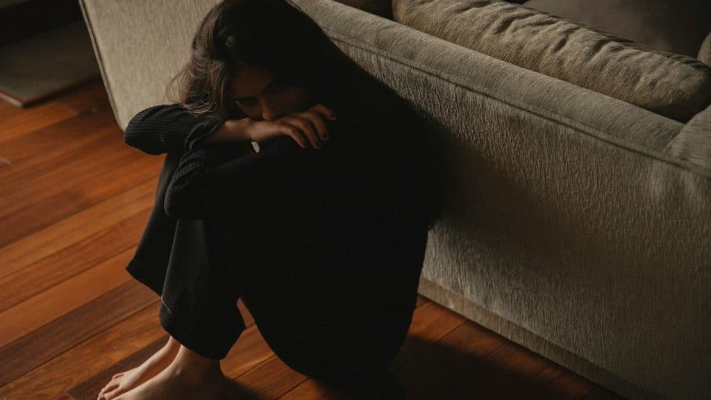 A sad woman sitting on the floor in a fetal position while leaning against a couch.