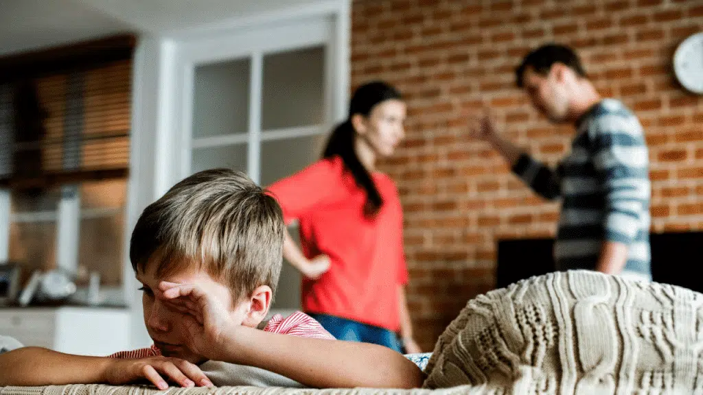 A young boy rubs his eye in the foreground while two adults argue behind him.