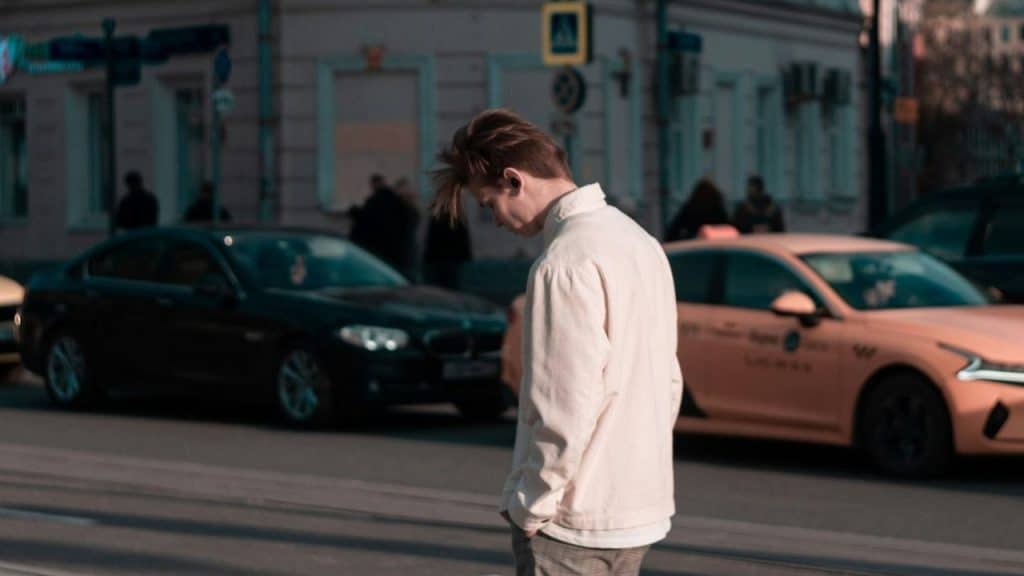 A man with light-colored hair walks across a city street with his head tilted downward.
