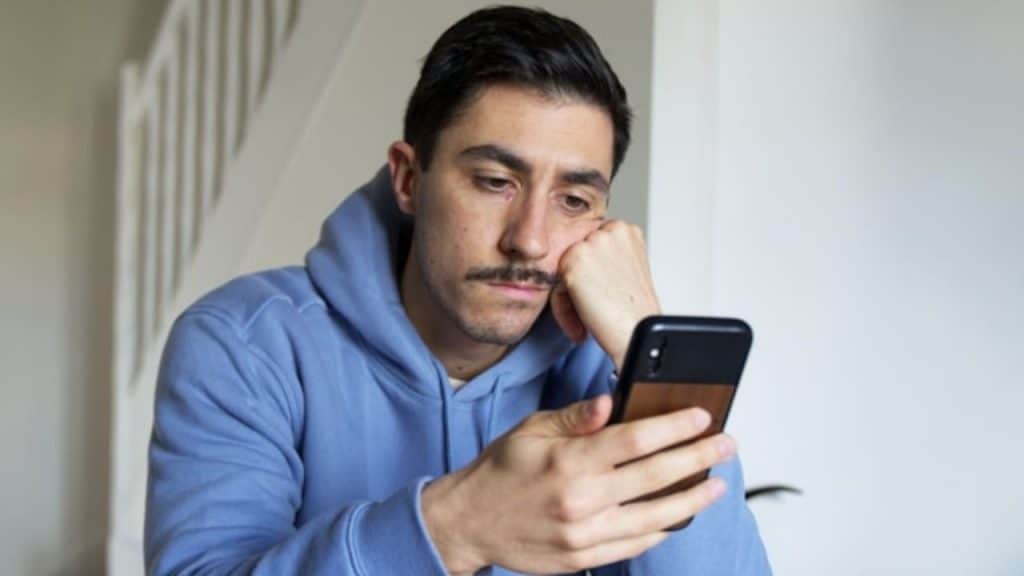 A man with a mustache rests his chin on his hand while looking at a smartphone.