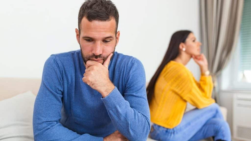 A man in a blue sweater sits in the foreground while a woman sits behind him.
